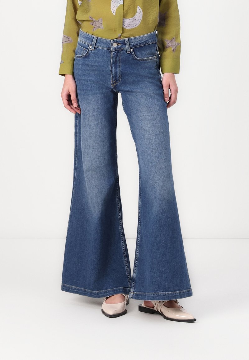 Jean bleu à jambes larges porté avec des chaussures slingback beiges à bout pointu et un chemisier vert olive avec des motifs de lune et d'étoiles.