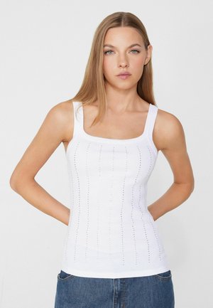 Giovane donna con lunghi capelli lisci indossa un top bianco senza maniche in maglia e jeans blu, in piedi davanti a uno sfondo bianco semplice.