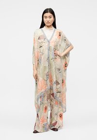 Γυναίκα που φορά διαφανές καφτάνι με λουλουδάτο σχέδιο σε απαλούς παστέλ τόνους, συνδυασμένο με παντελόνι φαρδιάς γραμμής στο ίδιο μοτίβο και σκούρα παπούτσια.