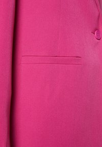 Pink blazer detail featuring a smooth texture, slim lapel, and a single chest pocket, complemented by a matching button.