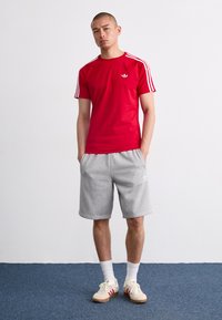 Piros, rövid ujjú póló, fehér háromsávos mintával a vállakon, szürke sorttal kombinálva. Fehér sportcipő piros részletekkel.