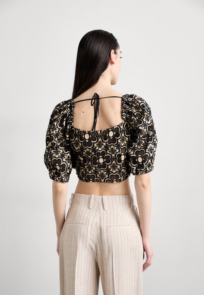 Black and gold floral crop top with puff sleeves and open back, featuring a tied detail at the neck and a textured fabric. Paired with striped beige pants.