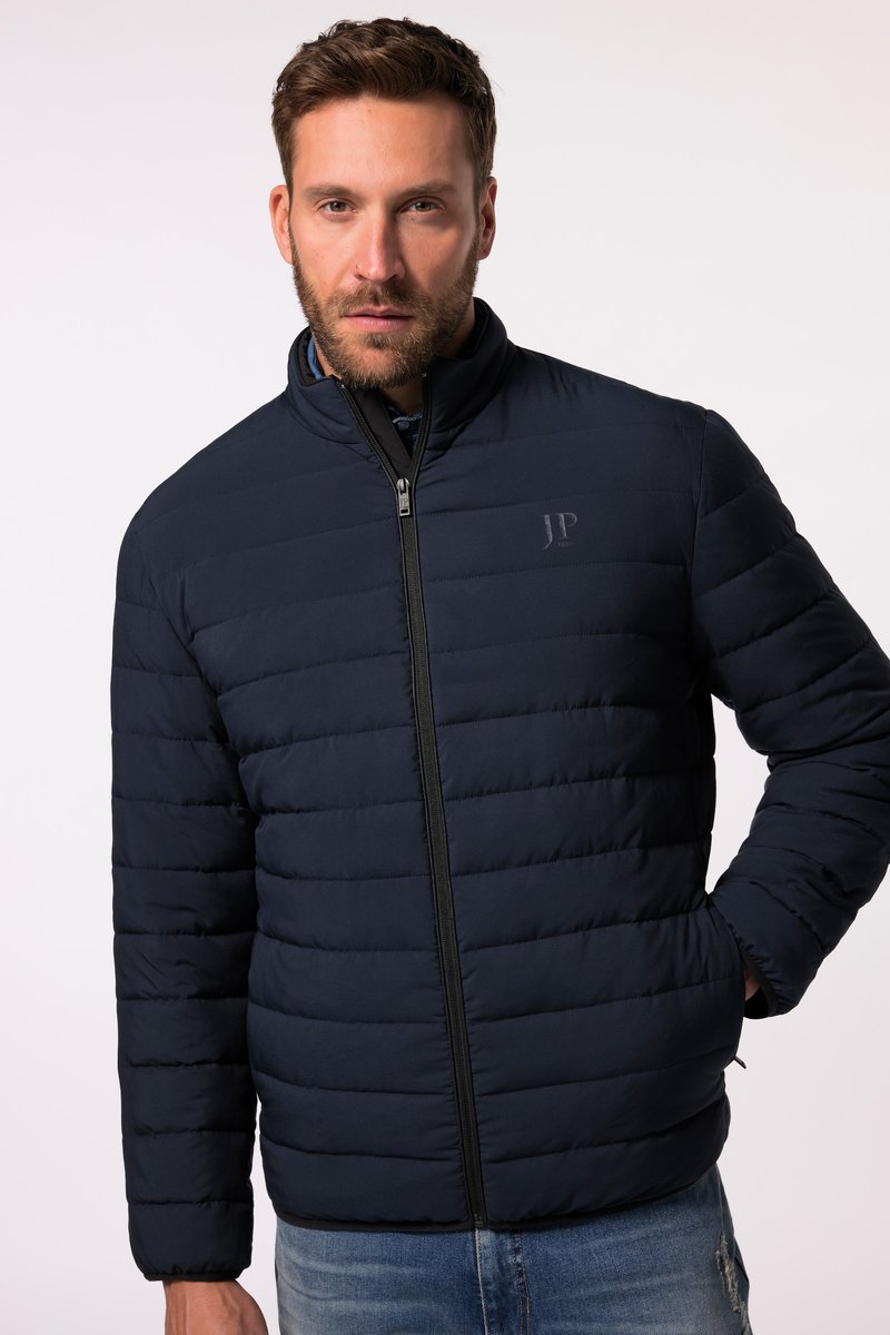 Mann trägt eine dunkelmarineblaue, gesteppte Jacke mit Reißverschluss und hohem Kragen sowie hellblaue Jeans, stehend vor einem einfarbigen Hintergrund.