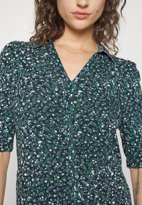 Femme aux cheveux bouclés portant un haut à fleurs vert, blanc et bleu marine avec un décolleté en V et des manches mi-longues, tatouage visible sur l'avant-bras droit.