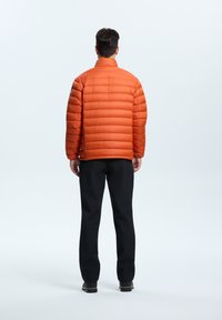 Orange pufferjakke med vertikal quiltedesign, høj krave og elastiske manchetter; parret med sorte bukser og robuste sorte støvler.