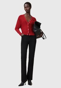 Cardigan rosso con scollatura a V profonda e quattro grandi bottoni, abbinato a pantaloni neri attillati e tacchi a punta neri, tenuto con una borsa nera.