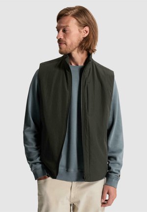 Mand med lyst brunt hår bærer en mørkegrøn ærmeløs vest over en blå langærmet skjorte og beige bukser, kigger til sin venstre side.