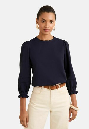 Blusa blu navy con maniche a sbuffo e polsini arricciati, con scollatura scalloppata e dettagli sottili. Abbinata a pantaloni di colore chiaro.