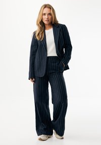 Granatowy garnitur w prążki z dopasowaną marynarką i spodniami o szerokich nogawkach, zestawiony z białym topem. Tkanina wydaje się mieć fakturę, do tego neutralne buty.