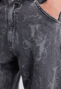 Pantalones vaqueros de mezclilla negra con un acabado desgastado, que presentan sutiles patrones gráficos. Diseñados con un estilo clásico de cinco bolsillos y detalles en metal.