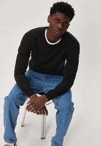 Black long-sleeve shirt with a white crewneck detail, paired with light blue denim jeans, seated on a metal chair.
