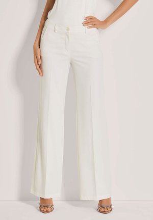 Frau in hochtaillierten, weißen, weit geschnittenen Hosen mit Gürtelschlaufen und praktischen Taschen, die auf einem schlichten hellen Hintergrund steht. Sie trägt Absatzsandalen.