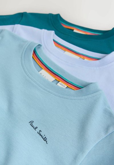 Tres camisetas apiladas: azul claro, verde azulado y lavanda. Cada una tiene cuello redondo, material suave de algodón y un detalle de cuello con rayas coloridas.