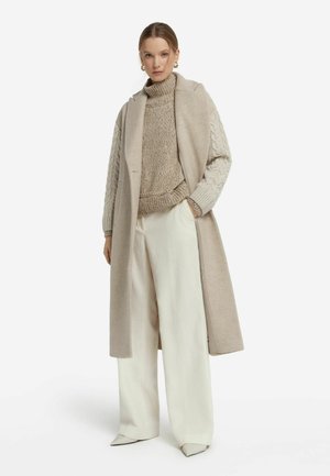 Beżowy długi płaszcz z wyprofilowanym kołnierzem, zestawiony z jasnobrązowym swetrem w warkocze i szerokimi białymi spodniami w prążki. Eleganckie czółenka na obcasie.