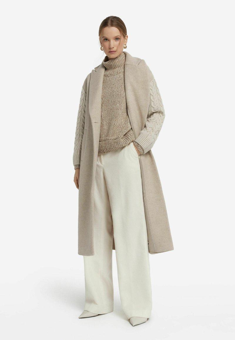 Beige lång kappa med strukturerad krage, i kombination med en ljusbrun kabelstickad tröja och vida vita ribbade byxor. Eleganta spetsiga klackar.