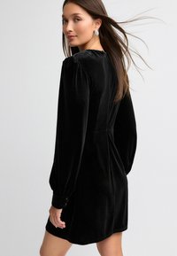 Schwarzes Samtkleid mit langen Puffärmel, tailliertem Schnitt und geknöpften Manschetten. Verfügt über einen dezenten Reißverschluss auf der Rückseite und hat eine reiche Textur.