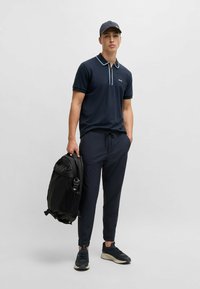 Polo shirt blu navy con colletto a contrasto, pantaloni scuri leggeri, zaino nero e sneakers blu navy, esposti su uno sfondo neutro.