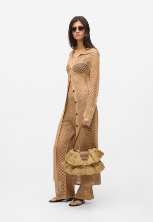 Mujer vistiendo un cárdigan largo de crochet beige y pantalones, con un sujetador negro visible debajo, sandalias negras, gafas de sol, sosteniendo un bolso beige con volantes.