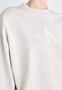 Jasno beżowa bluza z okrągłym dekoltem. Zdobiona haftowanym logo "Calvin Klein Jeans" w subtelnym, tonowanym wzorze. Miękka faktura materiału.