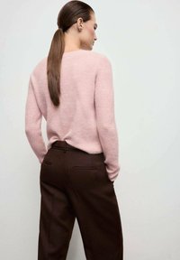 Donna con una lunga coda di cavallo marrone che indossa un maglione di maglia rosa chiaro e pantaloni marroni scuri a vita alta, in piedi con le mani in tasca.