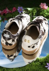 Paire de baskets blanches et noires à taches avec semelles marron portant l’inscription "CAVAL", posées sur une surface réfléchissante entourée de mousse verte et de fleurs colorées.
