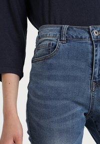 Jeans de mezclilla azul con una cintura de altura media, diseño de cinco bolsillos, desvanecimiento sutil y costuras de contraste en las costuras.