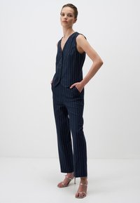 Marineblauw streepmotief vest en broek set, met een diepe V-halslijn, mouwloos ontwerp, twee zakken en een getailleerde pasvorm. Zilveren sandaaltjes met bandjes.