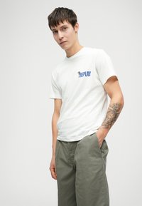 Jauns vīrietis ar īsiem tumšiem matiem, ģērbies baltā "REPLAY" T-kreklā un zaļās biksēs, ar sīki izstrādātu tetovējumu uz kreisā apakšdelma.