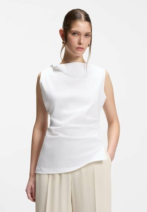 Jeune femme aux cheveux coiffés en arrière portant un haut blanc sans manches et un pantalon beige à jambes larges, sur un fond clair uni.