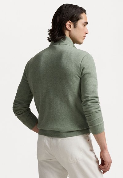 Green, lightweight sweater with a high collar and ribbed cuffs. Smooth texture, fitted design, worn with white pants.
