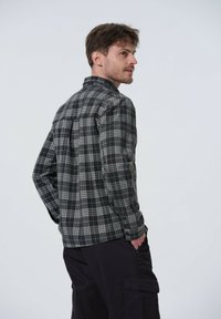 Musta ja harmaa ruudullinen flanellipaita pitkillä hihoilla, nappikauluksella ja takasaumalla, yhdistetty tummiin cargo-housuihin.