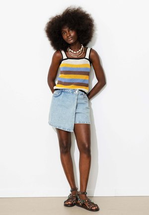 Femme avec une coiffure afro portant un débardeur rayé en maille, une jupe portefeuille en jean clair, des colliers coquillages superposés et des sandales noires cloutées, debout contre un mur.