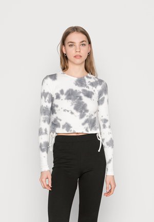 Top de mangas compridas cortado em tecido branco com padrão de tie-dye cinza, apresentando cordões de ajuste laterais e uma textura canelada. Combinado com calças pretas.