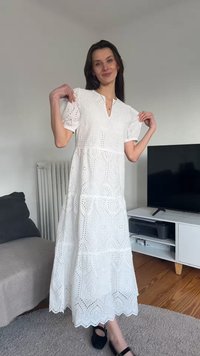Γυναίκα ποζάρει μέσα σε λευκό midi φόρεμα με δαντέλα και φουσκωτά μανίκια, φορώντας μαύρα επίπεδα παπούτσια, στέκεται κοντά σε τηλεόραση και καναπέ.