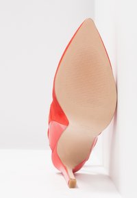 Chaussure à talon haut rouge reposant sur le côté, montrant une semelle beige lisse avec une prise texturée près du bout pointu.