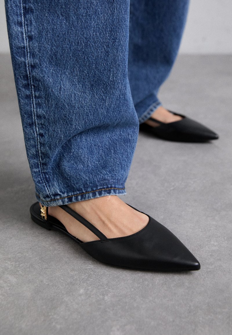 Melnas ādas norādītas slingback kurpes ar zemu papēdi, ar zeltainiem akcentiem, valkātas kopā ar zilām džinsu biksēm.