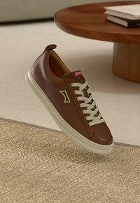 Brauner Ledersneaker mit weißen Schnürsenkeln, einem strukturierten braunen Abschnitt und einem Logodetail. Verfügt über eine cremefarbene Gummisohle und ein rotes Etikett.