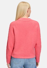 Blonde vrouw die wegkijkt, gekleed in een felroze gemêleerde trui en lichtblauwe denim jeans tegen een effen achtergrond.