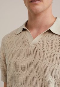 Beige knitted polo shirt with a V-neck and short sleeves, featuring a hexagonal pattern and a soft collar. Textured fabric with breathable design.