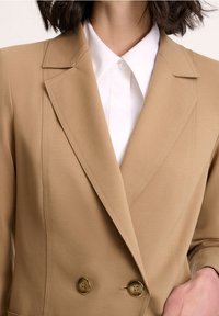 Beiger doppelreihiger Blazer mit abgesteppten Revers und goldenen Knöpfen. Hergestellt aus glattem Stoff mit präzisen Nähten und einer strukturierten Passform.