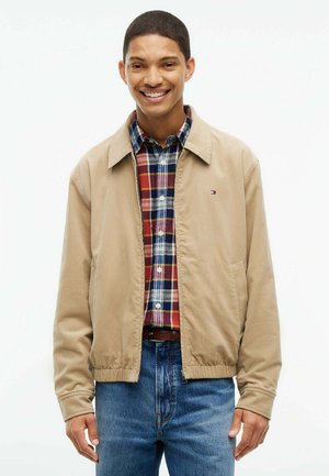 Ung mand smiler, iført en beige jakke over en rød og blå ternet skjorte og blå jeans, står foran en hvid baggrund.