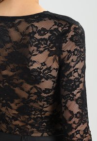 Haut en dentelle noire avec des motifs floraux, texture transparente, manches longues et encolure arrondie. Le design présente des détails complexes et une finition délicate.