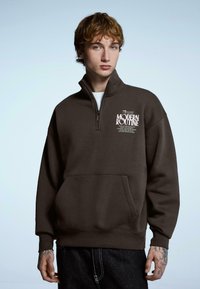 Ciemnobrązowa bluza z zamkiem na szyi, z przednią kieszenią typu kangurek, luźnym krojem, ściągaczami na rękawach i nadrukowanym napisem na piersi.