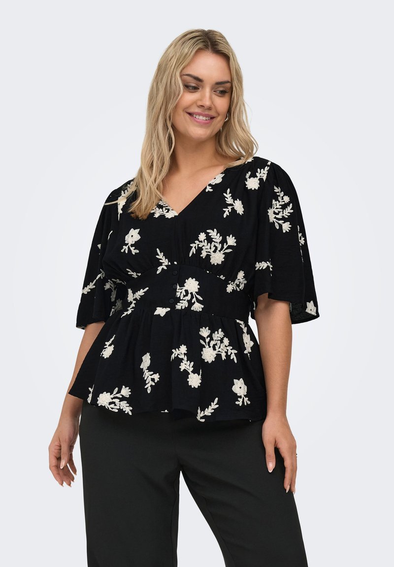 Blonde woman wearing black blouse with white floral embroidery, V-neck, short flutter sleeves, and black pants, smiling and looking down.