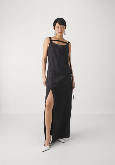 Rochie maxi din satin negru cu decolteu asimetric, fante pe lateral și detalii decorative la curea. Asortată cu pantofi cu vârf ascuțit albi.