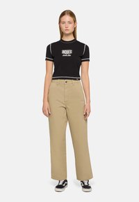 Zwart cropped t-shirt met witte accentstiksels en logo, gecombineerd met beige wijdlopende broek en zwarte sneakers.