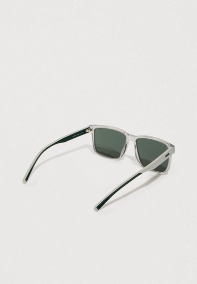 Lunettes de soleil à monture transparente avec des verres vert foncé, au design angulaire épuré, et des branches vert foncé contrastantes. Accents matériels minimalistes.