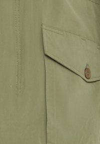 Jachetă khaki cu o textură netedă, două buzunare cu clapetă și un accent dintr-o buton maro. Materialul are o strălucire subtilă și cusături vizibile.