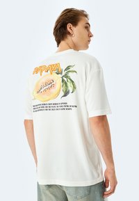 Hvid bomulds t-shirt med et farverigt papaya-grafik og teksten "PAPAYA" på ryggen. Korte ærmer, afslappet pasform og ribbestrikket halsudskæring.