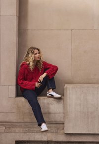 Red cable-knit sweater, oversized fit, paired with dark blue jeans and white sneakers. Textured, cozy look with a hood detail.